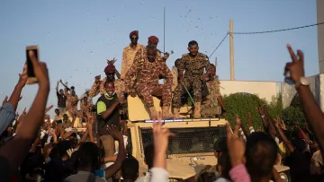 L'image montre un groupe de personnes en liesse rassemblées autour d'un véhicule militaire. Des soldats, vêtus d'uniformes camouflés, sont visibles en hauteur sur le véhicule, tandis que la foule en dessous lève les bras et semble manifester de la joie ou de l'enthousiasme. On peut apercevoir des confettis ou des éclats de couleur qui ajoutent à l'ambiance festive. L'atmosphère générale semble être celle d'une célébration ou d'un événement important.
