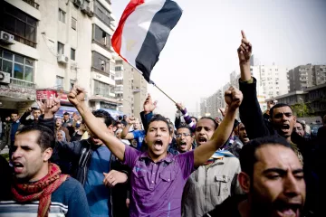 The image depicts a crowd of people in a street, likely engaged in a protest or demonstration. A man in the foreground is holding an Egyptian flag aloft and appears to be shouting passionately. The crowd around him is animated, with several individuals raising their fists and expressing strong emotions. The scene captures a sense of unity and determination among the protestors, amidst urban surroundings with buildings and visible signs of city life.