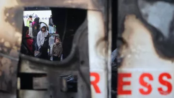 L'image montre un groupe de personnes, y compris des enfants, rassemblées dans un environnement urbain perturbé. On aperçoit une structure endommagée, peut-être un véhicule, qui présente des signes de destruction. Le reflet dans la structure suggère une atmosphère de tension ou de conflit, tandis que les visages des personnes expriment diverses émotions. L'environnement semble chaotique, soulignant une situation difficile.