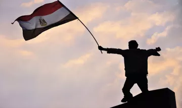 L'image montre une silhouette d'une personne debout sur une plateforme, levant un drapeau. Le drapeau est égyptien, avec des couleurs rouge, blanc, noir et le symbole du pays. L'arrière-plan présente un ciel avec des nuages aux teintes douces, créant une ambiance de détermination et de célébration. Cette scène évoque des sentiments de patriotisme et de protestation ou de victoire.