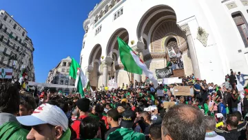 L'image montre une grande foule rassemblée devant un bâtiment avec une architecture traditionnelle. Les manifestants brandissent des drapeaux algériens et des pancartes, exprimant probablement des revendications politiques ou sociales. La scène est en plein air, sous un ciel bleu, et il y a une atmosphère de mobilisation collective, avec des personnes de tous âges vertes et rouges.