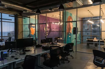 L'image montre un espace de bureau moderne avec de grandes fenêtres offrant une vue sur l'extérieur. Les murs sont décorés de peintures murales colorées représentant des visages stylisés. Le mobilier est minimaliste et fonctionnel, avec des tables de travail et des chaises ergonomiques. L'éclairage est tamisé, créant une ambiance chaleureuse malgré le design contemporain. On peut également voir des ordinateurs sur les bureaux, indiquant que l'espace est utilisé pour le travail.