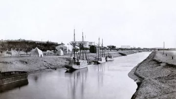 L'image montre une scène tranquille et nostalgique d'un canal ou d'une rivière. On y voit plusieurs bateaux amarrés, leur reflet se mêlant à l'eau calme. En arrière-plan, il y a des tentes et des structures qui évoquent une installation temporaire ou un camp. Le paysage environnant semble naturel, avec de la terre et des arbres. Cette photographie en noir et blanc donne une impression de sérénité et évoque une époque passée.