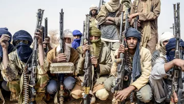 L'image montre un groupe d'hommes armés, probablement dans un environnement désertique. Ils portent des vêtements typiques de régions arides, comme des turbans et des foulards qui cachent partiellement leur visage. Chaque homme tient une arme à feu, et l'ambiance générale semble sérieuse et déterminée. Le groupe est organisé et se présente en ligne, ce qui suggère une certaine discipline. Le fond désertique renforce la dureté du cadre dans lequel ils se trouvent.
