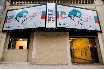L'immagine mostra un edificio con una facciata decorata da grandi manifesti. I manifesti annunciano un progetto artistico intitolato "The Restoration Project" di Youssef Chahine, con date di evento indicate dal 12 al 22 settembre. Si può notare un design grafico accattivante, con illustrazioni e testo in arabo e in inglese. La zona sembra essere un luogo di cultura o un centro espositivo, con elementi architettonici tipici di un contesto urbano.