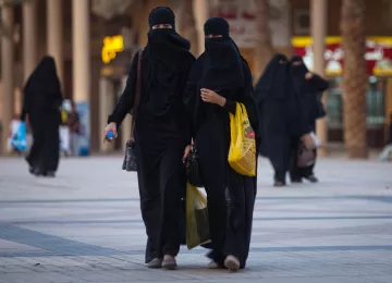 L'image montre deux femmes marchant côte à côte dans une zone urbaine. Elles portent des vêtements noirs, y compris des niqabs qui couvrent leur visage, laissant seulement les yeux visibles. L'environnement semble animé, avec des bâtiments en arrière-plan et d'autres personnes se déplaçant dans la même direction. L'une des femmes tient un sac, tandis que l'autre semble tenir un objet dans sa main. L'éclairage et l'architecture suggèrent un cadre contemporain.