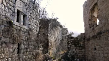 الصورة تظهر مشهدًا لموقع تاريخي قديم يحتوي على بقايا مبانٍ حجرية. نلاحظ جدرانًا متداعية وأجزاء مهدمة مع بعض النوافذ المفتوحة. الأرض محاطة بالأشجار والنباتات، ويبدو أن المنطقة مهجورة منذ فترة طويلة. الضوء الطبيعي ينعكس بشكل جميل على الحجر، مما يضفي جوًا هادئًا وغامضًا على المكان.