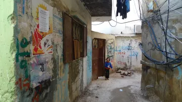 L'image montre une ruelle étroite et dégradée, bordée de murs en béton sur lesquels sont peints des graffitis et des affiches colorées. À gauche, une porte en bois est partiellement ouverte, tandis qu'à droite, on peut voir des fils électriques accrochés. Au fond de la ruelle, un jeune enfant se tient près de l'entrée, regardant vers l'objectif. L'atmosphère semble à la fois désolée et animée par les couleurs des murs et des affiches.