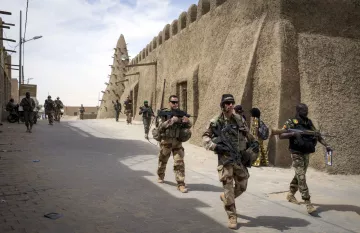The image depicts a group of soldiers walking through a narrow street in a fortified area. They are dressed in military gear and armed with rifles. The architecture features traditional, mud-brick structures, which create a historical and cultural backdrop. The scene conveys a sense of military presence and activity, with more soldiers seen in the background, indicating a coordinated operation or patrol. The atmosphere appears to be tense and vigilant, typical of a military environment.