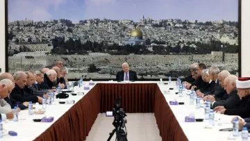 L'image montre une réunion se tenant autour d'une grande table ovale, avec plusieurs personnes assises de chaque côté. Au fond, il y a une grande image d'une ville, probablement Jérusalem, avec le dôme doré d'une mosquée bien visible. Les participants, qui semblent être des hommes âgés, portent des costumes sombres et affichent une expression sérieuse. Au centre de la table, une personne (probablement un chef ou un président) s'exprime pendant que les autres l'écoutent attentivement. Des bouteilles d'eau et des serviettes sont disposées sur la table. L'ambiance semble solennelle et attentive.
