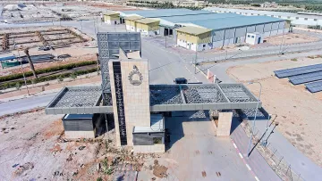 L'image montre un complexe industriel ou commercial avec une entrée bien marquée. On peut voir un portail moderne, probablement en métal et en pierre, qui semble être la pièce maîtresse de l'entrée. À l'arrière plan, il y a plusieurs bâtiments de différentes tailles, dont un plus grand qui pourrait servir de site de production ou d'entrepôt. Le terrain environnant est désertique,avec quelques infrastructures comme des panneaux solaires. En arrière-plan, on distingue un nuage de fumée, suggérant une activité ou un incendie à proximité.