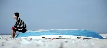 L'image montre un jeune garçon assis sur une coque de bateau échouée sur une plage. Il porte un t-shirt à rayures et un pantalon rouge. L'environnement est clair, avec un ciel bleu pâle et une plage de sable. Le garçon semble pensif, tourné vers l'horizon, tandis que la coque du bateau, abandonnée et usée, ajoute un élément de mélancolie à la scène.