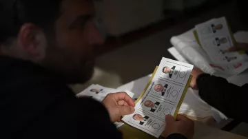 در تصویر، فردی دیده می‌شود که در حال بررسی یک برگه است. این برگه شامل تصاویری از چند نفر با نام‌ها و اطلاعات مرتبط با آن‌هاست. احتمالاً این برگه به انتخابات یا رأی‌گیری مربوط می‌شود. فرد در حال تأمل است و در محیطی شبیه به یک مرکز رأی‌گیری قرار دارد. نور به آرامی به صحنه می‌تابد و بر تمام جزئیات تأثیر می‌گذارد.