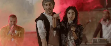L'image montre des artistes en pleine performance musicale, avec une ambiance vibrante et colorée. Au centre, une femme chante dans un microphone, accompagnée d'un homme à ses côtés qui semble participer activement à la performance. En arrière-plan, d'autres musiciens se joignent à l'animation, entourés de lumière et de fumée, créant une atmosphère dynamique et festive. Les expressions des artistes indiquent de l'enthousiasme et de l'énergie.