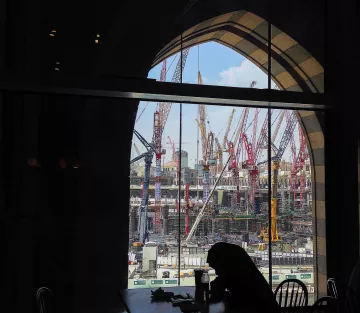 L'image montre un chantier de construction vu à travers une grande fenêtre à arc. À l'intérieur, on aperçoit une silhouette assise à une table. De l'autre côté de la vitre, de nombreux grues et engins de chantier se dressent, travaillant à un projet de grande envergure. Le ciel est partiellement dégagé, ajoutant une touche de lumière à la scène. L'ensemble dégage une atmosphère de travail intensif et de développement urbain.