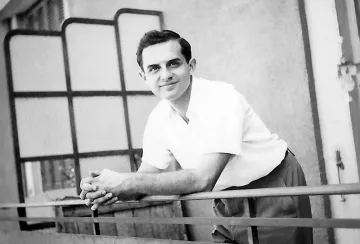 در این تصویر، مردی جوان به نظر می‌رسد که در حال لبخند زدن است. او با آستین‌های کوتاه و پیراهن سفیدی لباس پوشیده و در بالکن یا جایی مشابه ایستاده است. دستانش را بر روی نرده‌های بالکن گذاشته و به دوربین نگاه می‌کند. پس‌زمینه تصویر شامل دیواری ساده و پنجره‌ای با شیشه‌های مشبک است. جو تصویر دوستانه و صمیمانه به نظر می‌رسد.