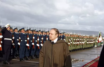 L'image montre un homme en manteau se tenant devant un alignement de soldats en uniforme. Les soldats, majestueux et bien rangés, saluent. L'arrière-plan est nuageux, créant une atmosphère cérémonielle. Le sol semble humide, indiquant peut-être qu'il a plu récemment. L'homme au premier plan a une expression pensive, ajoutant un ton sérieux à la scène.