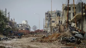 الصورة تظهر مشهدًا لمدينة مدمرة، حيث تبدو المباني متضررة بشدة، مع بقايا الهياكل المعدنية وأجزاء من الأنقاض. الطريق في المنتصف مليء بالحصى والأحجار، ويبدو غارقًا في الخراب. يظهر في الخلفية بعض الأعمدة الكهربائية التي لم تعد تعمل، مما يضيف إلى الجو القاتم والموحش للمكان. الأجواء عامة غائمة، مما يزيد من شعور الخواء واليدانة في هذا المشهد.