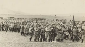 در این تصویر، گروهی از سربازان در یک صف منظم ایستاده‌اند. آنها uniform نظامی پوشیده‌اند که شامل لباس‌هایی با رنگ‌های خاکی و کلاه‌های مخصوص است. برخی از آن‌ها در حال حمل سلاح هستند و عده‌ای دیگر به حالت آماده به کار ایستاده‌اند. پس‌زمینه تصویر، زمین وسیعی را نشان می‌دهد که در آن تعدادی دیگر از سربازان نیز دیده می‌شوند. این تصویر به نظر می‌رسد که مربوط به یک دوره تاریخی خاص باشد.