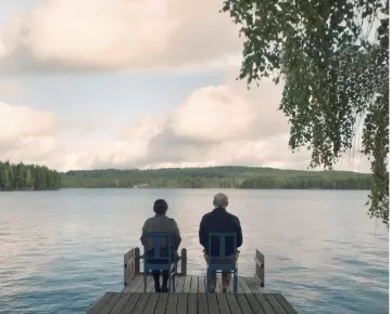 L'image montre deux personnes assises sur des chaises en bois sur une jetée qui s'avance dans un lac paisible. Ils regardent vers l'horizon, avec des arbres verdoyants en arrière-plan et un ciel nuageux au-dessus. L'atmosphère est calme et sereine, évoquant un moment de contemplation ou de discussion tranquille.