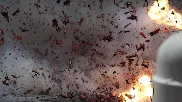 The image depicts an intense explosion, with fragments and debris scattered throughout the scene. There are bright flashes of light indicating the force of the blast, and the background appears smoky and chaotic, suggesting a highly energetic event. The overall atmosphere conveys a sense of destruction and turmoil.