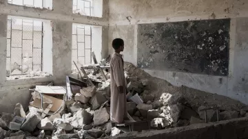 در این تصویر، یک پسر جوان در یک کلاس درس ویران شده ایستاده است. دیوارها و سقف کلاس آسیب دید‌ه‌اند و بخش‌هایی از زیرساخت‌ها به طور کامل تخریب شده‌اند. در اطراف او، زباله‌ها و آوارهای ساختمانی وجود دارد. یک تابلو سیاه با نوشته‌هایی بر روی دیوار وجود دارد، اما وضعیت آن نیز نامناسب است. نور از پنجره‌های شکسته به فضای داخلی می‌تابد و حس ناامیدی و ویرانی را منتقل می‌کند.