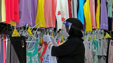 The image depicts a person wearing a black hijab and a face shield, shopping for clothing in a store. They are holding a white shopping bag and carefully examining different tops displayed on hangers. The store features a colorful array of clothing items, including various shades of purple, yellow, and floral patterns, hanging on racks in the background. The environment suggests a retail space focused on apparel, likely catering to a diverse range of styles.