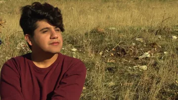 L'image montre une personne assise dans un champ de herbes hautes. La personne a les cheveux foncés et bouclés, et elle semble pensive, regardant au loin. Elle porte un pull de couleur bordeaux. L'environnement est naturel, avec une lumière douce, ce qui crée une atmosphère tranquille.