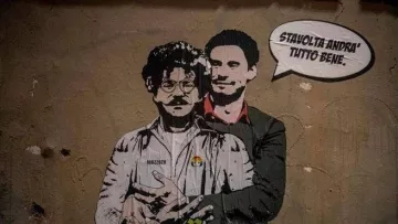 L'immagine mostra un murale che ritrae due figure. Una di esse ha un aspetto distintivo, con occhiali e baffi, mentre l'altra indossa un completo elegante. Sullo sfondo, si può notare un muro di colore neutro, e sopra le figure è presente un testo in italiano che dice "Stai tranquillo, andrà tutto bene". Inoltre, nella parte inferiore della figura è scritta la parola "حرية" (che significa "libertà" in arabo). L'opera sembra trasmettere un messaggio di speranza e solidarietà.