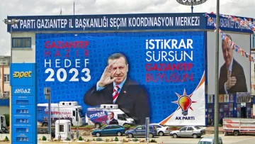 L'image montre un grand panneau publicitaire avec des slogans politiques. On y voit des phrases comme "GAZİANTEP HAZIR 2023" et "İSTİKRAR SÜRSÜN GAZİANTEP BÜYÜSÜN", suggérant une campagne électorale pour les élections de 2023. L'image présente également une figure politique qui salue, ajoutant une dimension dynamique à la publicité. Le fond inclut des symboles ou logos associés à un parti, probablement le parti AK. La scène est typique des campagnes électorales visuelles, avec une mise en avant forte du message et de la personnalité politique.