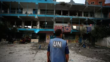 در این تصویر، یک مرد در لباس یونیفورم با نشانه "UN" (سازمان ملل) ایستاده است و به سمت یک ساختمان ویران شده نگاه می‌کند. ساختمان دارای دیوارهایی با رنگ‌های مختلف و برخی آسیب‌ها و خرابی‌ها است. در محیط اطراف، آثاری از ویرانی و خرابی مشاهده می‌شود و نیز تعدادی افراد دیگر در پس‌زمینه قرار دارند. فضای کلی تصویر حس ناراحتی و بحران را منتقل می‌کند.