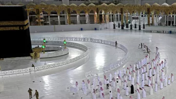 الصورة تُظهر الحرم المكي ومكة المكرمة، حيث يمكن رؤية الكعبة المشرفة في وسط الصورة. هناك عدد من المصلين يقفون في صفوف مرتبة لأداء الصلاة، ويرتدون الملابس التقليدية. الساحة المحيطة بالكعبة نظيفة وواسعة، مع وجود حواجز حول مناطق معينة. كما يظهر بعض رجال الأمن في الصورة، مما يدل على تنظيم المكان. تمثل هذه اللقطة أجواء العبادة والتقرب إلى الله، وخاصة في الأوقات التي تكون فيها أعداد المصلين محدودة.