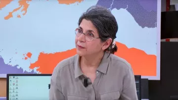 این تصویر فردی را نشان می‌دهد که در یک محیط خبری یا استودیو قرار دارد. او دارای موهای خاکستری و آرایش ساده است و عینک دارد. لباس او رسمی و به رنگ ملایم است. پس‌زمینه تصویر نقشه‌ای از جهان یا بخش‌هایی از آن را نمایش می‌دهد که به نظر می‌رسد به موضوع خبر مربوط می‌شود. چهره این فرد نشان‌دهنده توجه و تمرکز است.
