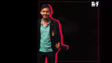 L'image montre un homme souriant, portant une chemise turquoise sous un veston noir. Il a une carte d'identification accrochée à son cou et tient un appareil photo à la main. Le fond est principalement noir, avec un contour rose qui souligne la silhouette de l'homme. En haut à droite, le logo "RSF" est visible.