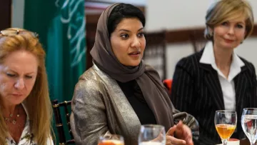 L'image montre un groupe de femmes assises autour d'une table, en train de discuter. Une femme au centre porte un hijab et un vêtement aux couleurs neutres, semblant engagée dans la conversation. À sa gauche, une autre femme aux cheveux longs et blonds, légèrement penchée, écoute attentivement. À droite, une femme en tailleur noir et blanc observe également. Sur la table, on peut voir des verres de boisson et de la vaisselle, avec des éléments de nourriture. L'environnement semble formel, propice à une réunion ou un déjeuner d'affaires.