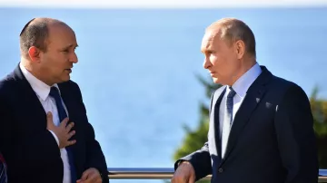 The image shows two men engaged in conversation by a seaside. One man is wearing a suit and has some form of head covering, indicating he may be of Jewish faith. The other man is also dressed in a suit and appears to be listening intently. They are both standing outdoors with a view of the water in the background. The setting suggests a formal or diplomatic meeting.