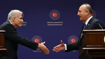 The image depicts two men engaged in a handshake, likely at a diplomatic or formal meeting. They are standing on either side of a wooden podium, with a background featuring the logo of the Turkish Ministry of Foreign Affairs. One man appears to be offering his hand in a gesture of greeting, while the other is responding. Both are dressed in formal attire, suggesting a professional context.