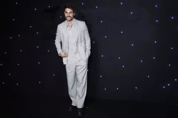L'image montre un homme portant un costume léger à rayures, composé d'une veste et d'un pantalon ample. Il a une moustache et un sourire, et se tient debout avec une pose décontractée. L'arrière-plan est sombre, parsemé de petites lumières blanches qui ajoutent un effet élégant à la scène.