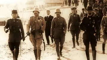 The image depicts a historical scene with a group of military personnel walking together in a line. They are dressed in formal military uniforms and accessories, including hats and medals, indicating a high rank or importance. The setting appears to be outdoors, possibly on a dirt or gravel path, with tents in the background suggesting a military camp. The overall tone of the photograph has a sepia or monochrome effect, conveying a sense of history. The individuals appear to be engaged in conversation or in a social setting, reflecting camaraderie among them.