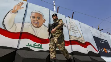 L'image montre un soldat en uniforme, armé et en marche, devant un mur peint. Sur le mur, il y a une fresque représentant un homme souriant et levant la main, probablement symbolisant une figure importante ou un leader. Le fond de la fresque inclut des éléments décoratifs et des couleurs qui évoquent le drapeau irakien. L'environnement semble être une zone urbaine, marquée par des installations de sécurité.