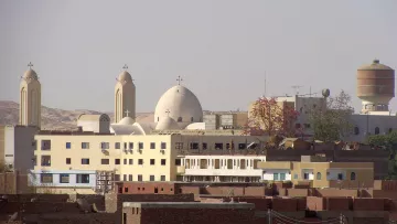 L'image montre un paysage urbain avec des bâtiments variés. On peut distinguer plusieurs églises avec des croix sur leurs dômes, caractéristiques de l'architecture religieuse. Des structures modernes cohabitent avec des bâtiments plus traditionnels, créant un contraste dans l'architecture. À l'arrière-plan, des collines ou des montagnes sont visibles, ajoutant une dimension naturelle au paysage. L'ensemble dégage une atmosphère de vie quotidienne dans une région marquée par son héritage culturel.