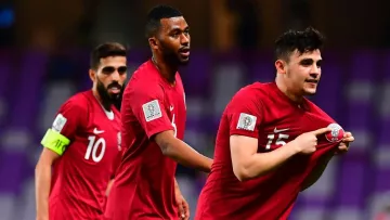 L'image montre des joueurs de football célébrant un moment de victoire ou d'accomplissement sur le terrain. Ils portent des maillots rouges avec des logos de l'équipe, et l'un d'eux semble montrer un symbole ou une médaille en soulevant son maillot. Les autres joueurs réagissent joyeusement derrière lui, montrant l'esprit d'équipe et la camaraderie. L'arrière-plan fait référence à un stade, créant une atmosphère d'événement sportif.