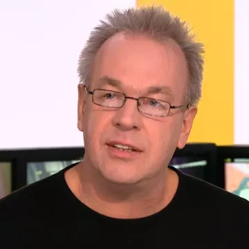 L'image montre un homme avec des cheveux gris et des lunettes. Il porte un t-shirt noir et semble en train de parler, avec un fond flou qui suggère un environnement de discussion ou d'interview.