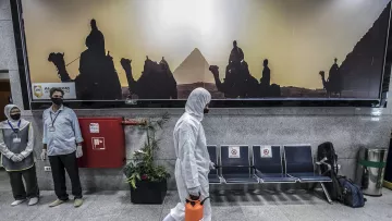 تظهر في الصورة مشهداً داخل محطة أو مطار. في الخلفية يوجد جدار مزين بصورة لأهرامات مصر مع silhouettes لحيوانات وحشود. في المقدمة، يسير شخص يرتدي بدلة واقية وقناع، حاملاً حقيبة برتقالية. بجانبه، يوجد موظفان يرتديان كمامات ويتوليان خدمات الاستقبال. في الزاوية، توجد مقاعد فائضة وبعض النباتات. تعكس الصورة أجواء احترازية مع لمسة ثقافية.