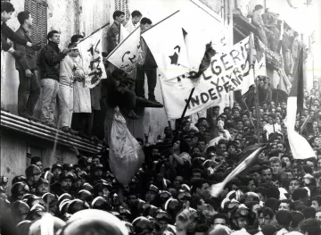 L'image montre une grande foule rassemblée, avec des individus brandissant des drapeaux et des bannières. L'atmosphère semble tendue et chargeée d'émotion, probablement liée à un événement historique. Des personnes se tiennent sur un bâtiment, tandis que d'autres sont regroupées en bas, certaines portant des uniformes, ce qui pourrait indiquer une présence policière ou militaire. Le message sur les bannières évoque des thèmes d'indépendance, suggérant un contexte de luttes sociales ou politiques.