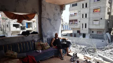 L'immagine mostra un uomo seduto su un divano in un ambiente che appare distrutto. Le pareti sono danneggiate e si possono vedere segni evidenti di distruzione. Nella stanza ci sono alcuni oggetti sparsi, come pezzi di ceramica e materiali vari. Alle spalle dell'uomo, le finestre danno su un paesaggio urbano suggerente di devastazione, con edifici danneggiati e in rovina visibili all'esterno. L'atmosfera è triste e riflette una situazione di difficoltà e lotta.