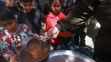 در این تصویر، گروهی از کودکان در حال دریافت غذا هستند. یکی از کودکان که لباس‌های رنگارنگ دارد، با دقت در حال ریختن خورش یا سوپ به ظرفش است. در پس‌زمینه، سایر کودکان نیز منتظر هستند تا نوبتشان برسد. محیط بیرونی به نظر شلوغ و غیررسمی است و نشان‌دهنده یک فعالیت جمعی برای سرو غذا به افراد نیازمند می‌باشد.