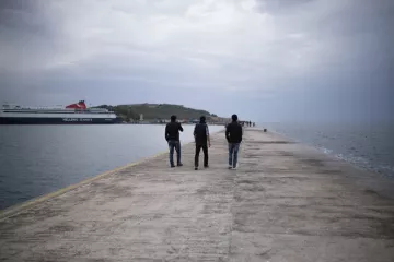 L'image montre trois personnes marchant sur un quai. Le ciel est nuageux, et l'atmosphère est calme. À l'arrière-plan, on peut voir un ferry amarré et une petite île. Le quai est en béton, et l'eau semble calme, reflétant les nuages. Les silhouettes des gens ajoutent une touche de vie à cette scène paisible.