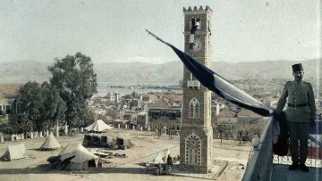 L'image montre une vue panoramique d'une place animée avec un grand bâtiment en pierre qui pourrait être une horloge ou une tour emblématique. À proximité, un soldat en uniforme se tient près de la balustrade, observant la scène. On peut voir des tentes disposées sur la place, peut-être pour un camp militaire ou une exposition. En arrière-plan, on distingue des collines et un plan d'eau, ajoutant à la profondeur du paysage. Le ciel semble dégagé, créant une atmosphère paisible.