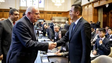 La imagen muestra a un grupo de personas en un entorno de reunión, probablemente en una conferencia o cumbre. En primer plano, dos hombres se están estrechando la mano, lo que sugiere un saludo cordial o un acuerdo. Uno de ellos tiene el cabello canoso y viste un traje oscuro, mientras que el otro es más joven y usa un traje también oscuro. Al fondo, se pueden ver otras personas sentadas alrededor de una mesa, con documentos y laptops, observando la interacción. El ambiente es formal y parece que se trata de un encuentro importante.
